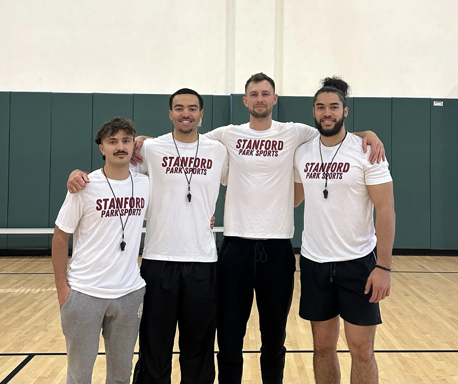 Four Stanford Park Sports coaches in matching shirts and whistles in a school gym
