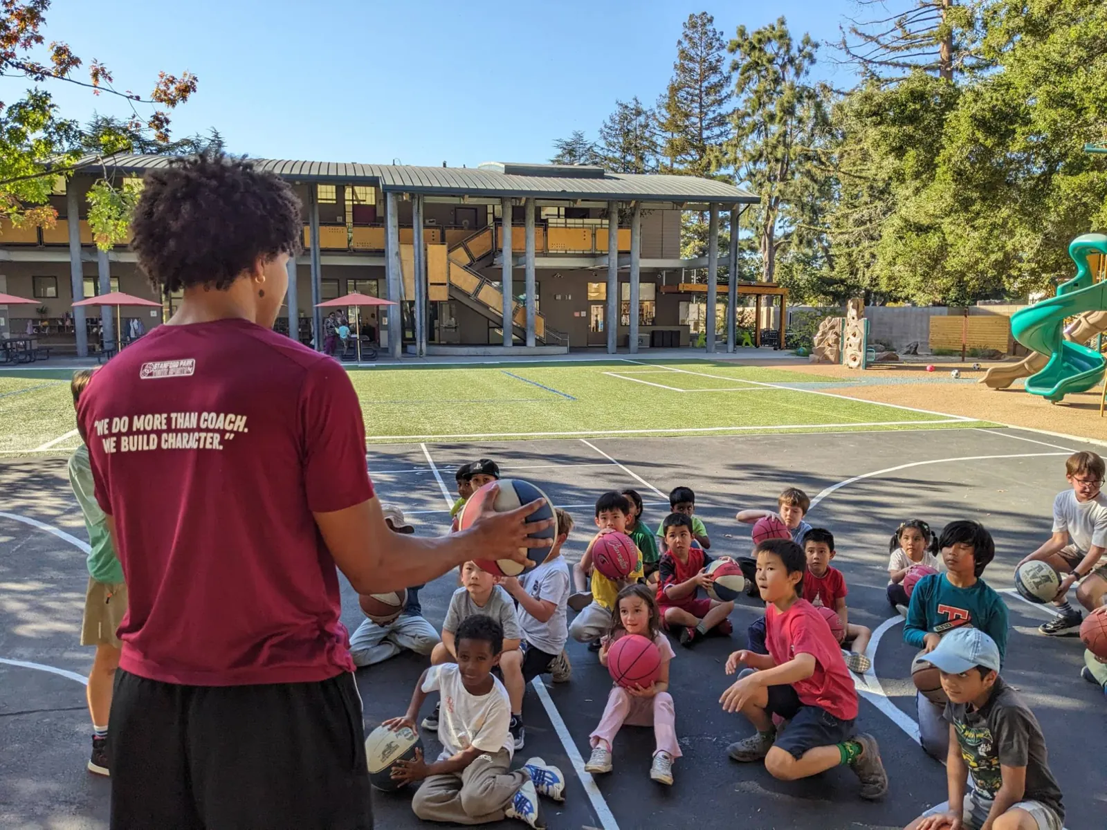 Stanford Park Sports coach in a shirt reading We do more than coach. We build character, leading a group of students with basketballs on a school court